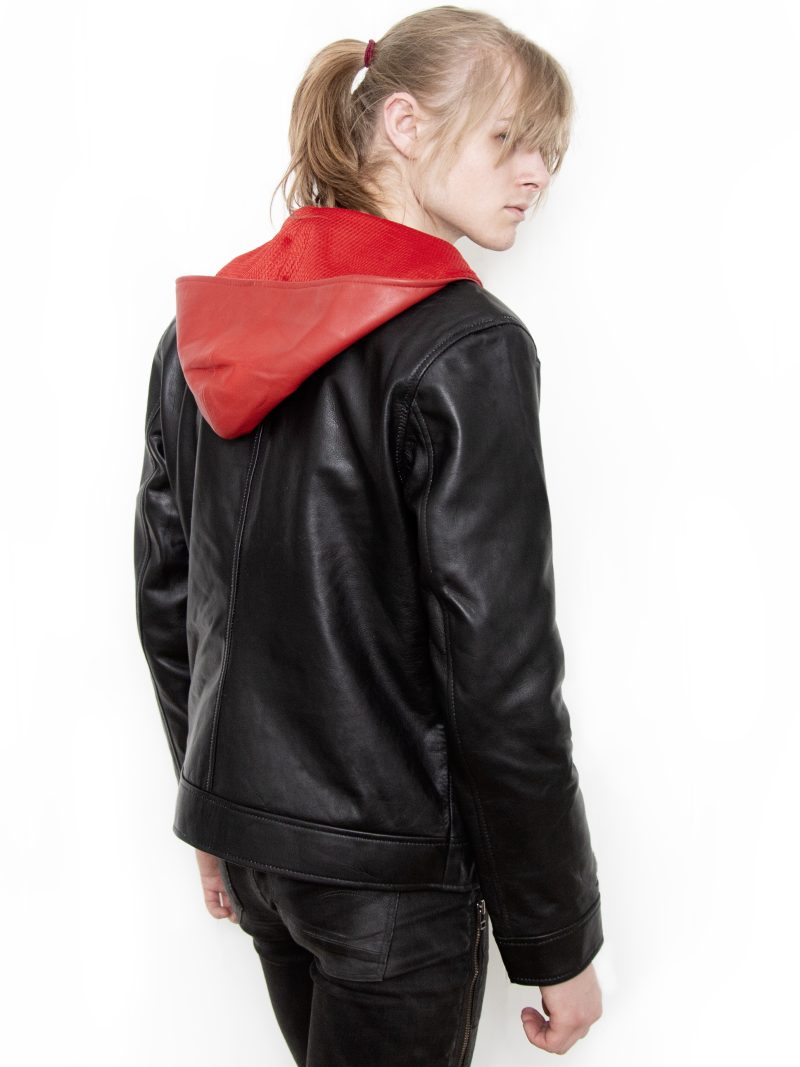 (Front): Photo of a model wearing a black leather jacket. The jacket is a motorcycle cut with a red hood and a red leather lining that is visible inside the cuffs and the folded down collar. There is also a strip of red leather either side of the front zipper. Where breast pockets typically would be on a jacket, there are [handpainted/patched] green eyes surrounded by white details. The collar has metal pop buttons to hold it in place. (Back): Photo of the same black leather jacket with a red hood. The jacket is a motorcycle cut. The inside of the hood has a mesh lining.