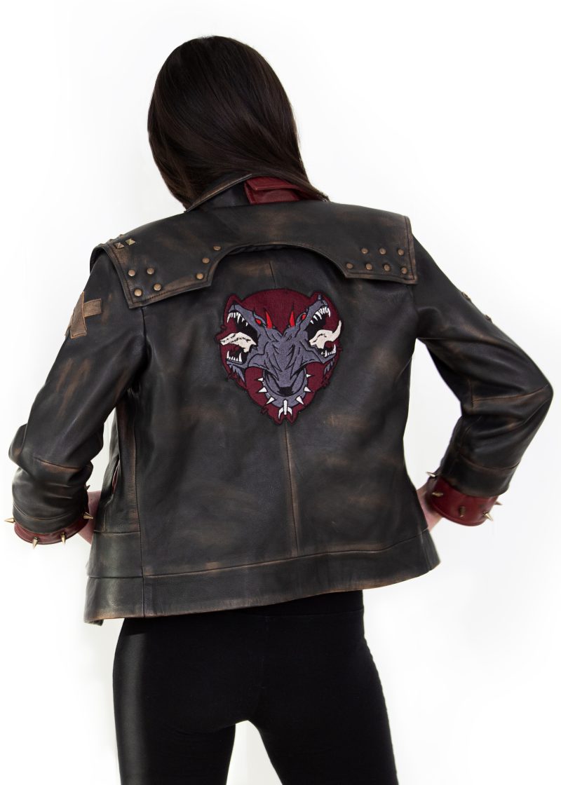 Photo of a model wearing a dark brown leather jacket. The back of the jacket is visible, showing the same 3/4 length red studded leather cuffs and the light brown X on the left upper arm. The back of the jacket has a large patch, showing a two-headed dog wearing a studded collar, both heads with their mouths open and teeth bared. The background of the patch is in the shape of an upside-down teardrop.The jacket pictured is the Vi arcane season 2 jacket.