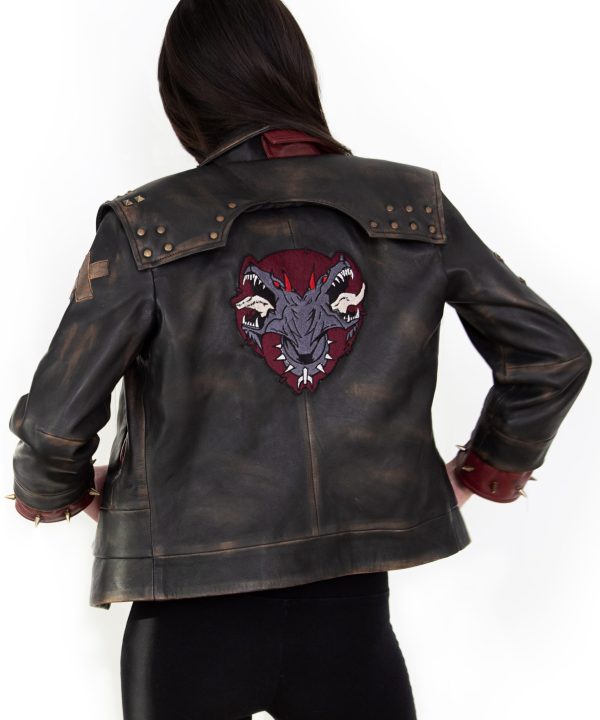 Photo of a model wearing a dark brown leather jacket. The back of the jacket is visible, showing the same 3/4 length red studded leather cuffs and the light brown X on the left upper arm. The back of the jacket has a large patch, showing a two-headed dog wearing a studded collar, both heads with their mouths open and teeth bared. The background of the patch is in the shape of an upside-down teardrop.The jacket pictured is the Vi arcane season 2 jacket.