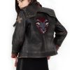 Photo of a model wearing a dark brown leather jacket. The back of the jacket is visible, showing the same 3/4 length red studded leather cuffs and the light brown X on the left upper arm. The back of the jacket has a large patch, showing a two-headed dog wearing a studded collar, both heads with their mouths open and teeth bared. The background of the patch is in the shape of an upside-down teardrop.The jacket pictured is the Vi arcane season 2 jacket.