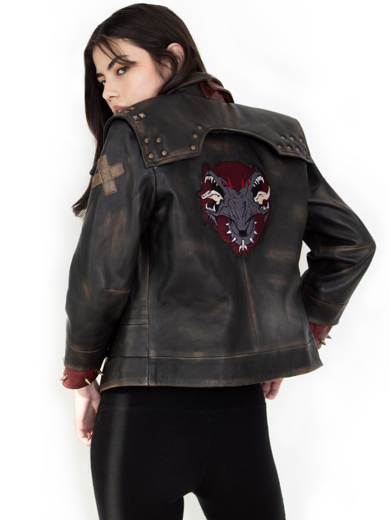 Photo of a model wearing a dark brown leather jacket. The back of the jacket is visible, showing the same 3/4 length red studded leather cuffs and the light brown X on the left upper arm. The back of the jacket has a large patch, showing a two-headed dog wearing a studded collar, both heads with their mouths open and teeth bared. The background of the patch is in the shape of an upside-down teardrop.The jacket pictured is the Vi arcane season 2 jacket.