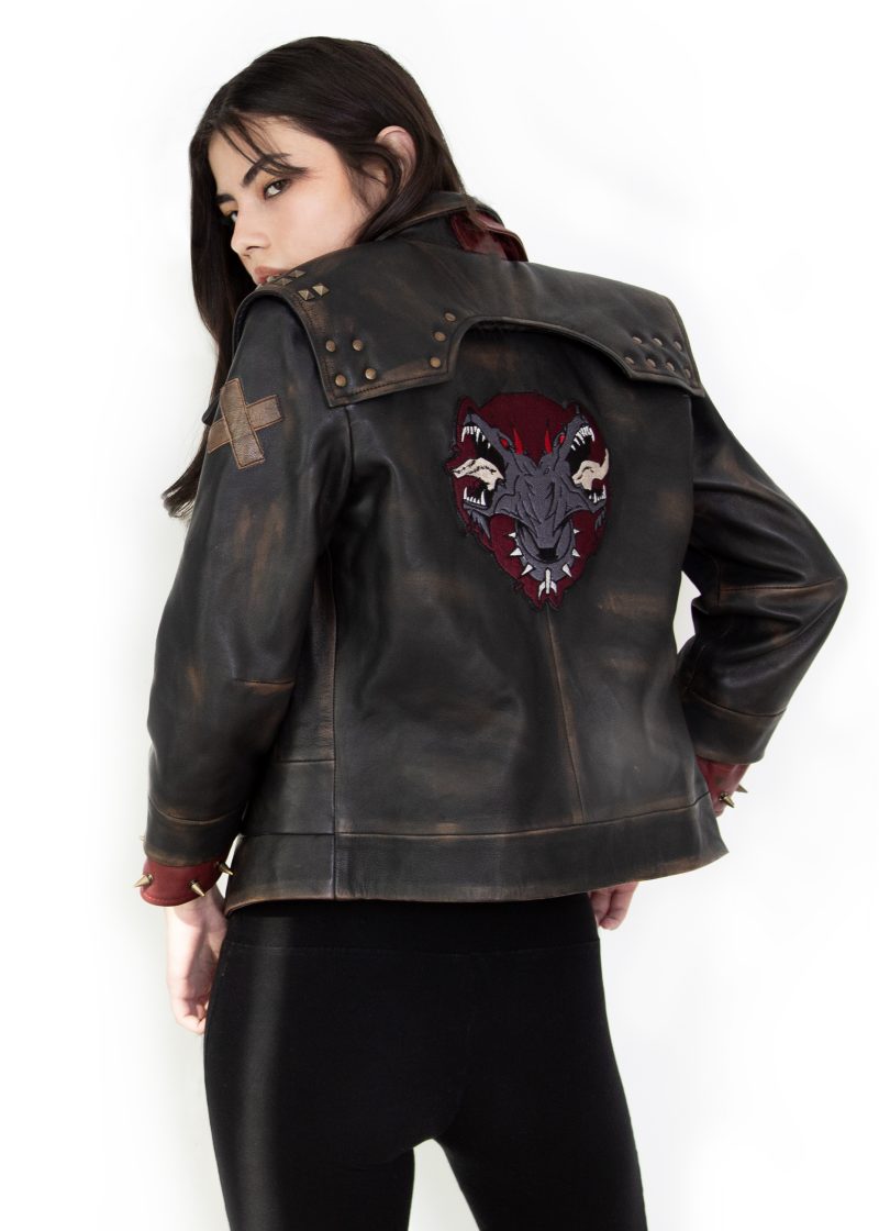 Photo of a model wearing a dark brown leather jacket. The back of the jacket is visible, showing the same 3/4 length red studded leather cuffs and the light brown X on the left upper arm. The back of the jacket has a large patch, showing a two-headed dog wearing a studded collar, both heads with their mouths open and teeth bared. The background of the patch is in the shape of an upside-down teardrop.The jacket pictured is the Vi arcane season 2 jacket.
